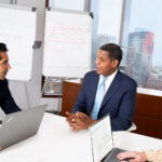 man in black suit conferring with financial consultant in blue suit in conference room with woman on laptop